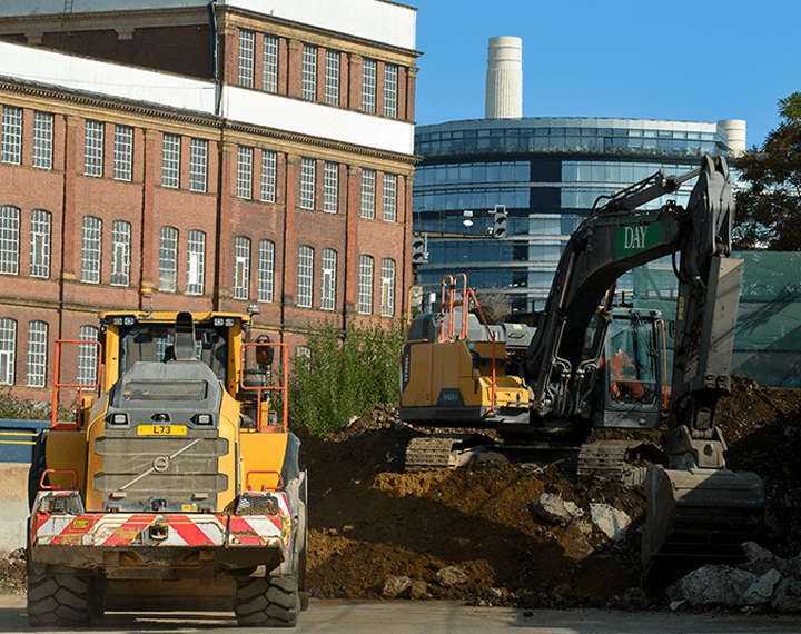 Yellow loading shovel and excavator next to a pile or rubble with buildings in the background