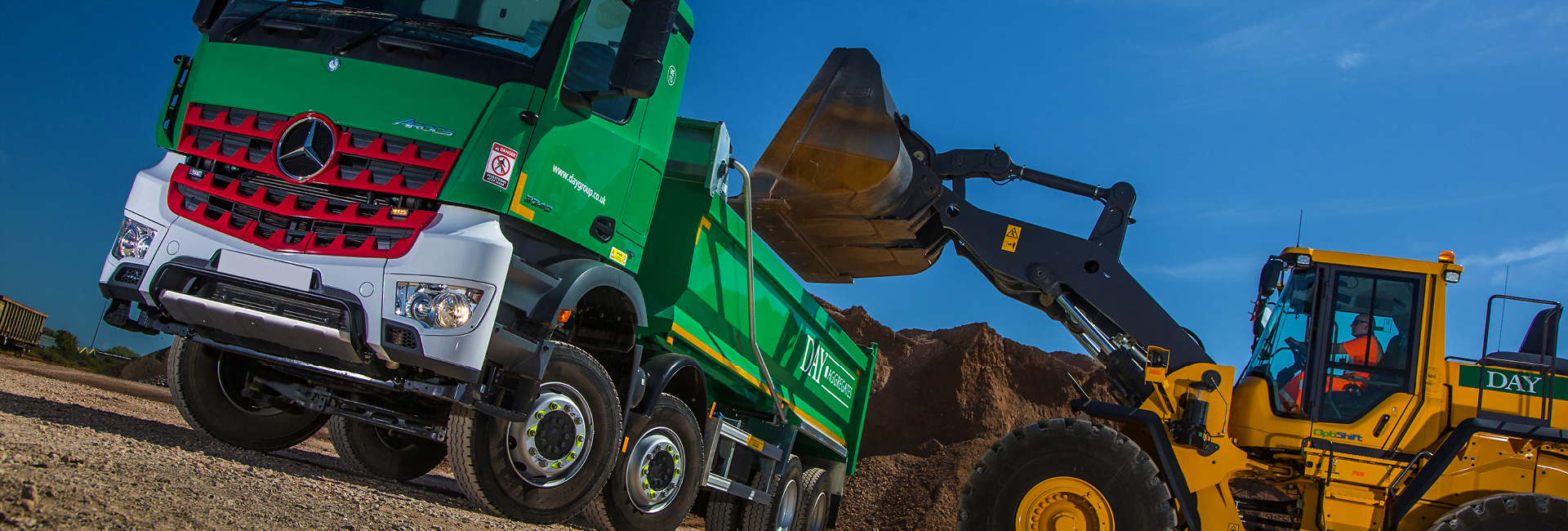 angled photo of yellow shovel tipping material into a green Day aggregates 8 wheeler lorry